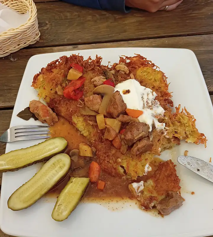 Placek po węgiersku z gulaszem, śmietaną i ogórkami kiszonymi na białym talerzu, na drewnianym stole